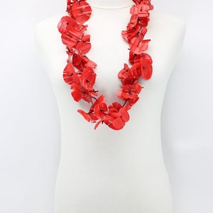 May include: A vibrant red floral necklace displayed on a white mannequin. The necklace is composed of numerous red flower-shaped elements, creating a layered and eye-catching design. The bright red color contrasts with the white background.