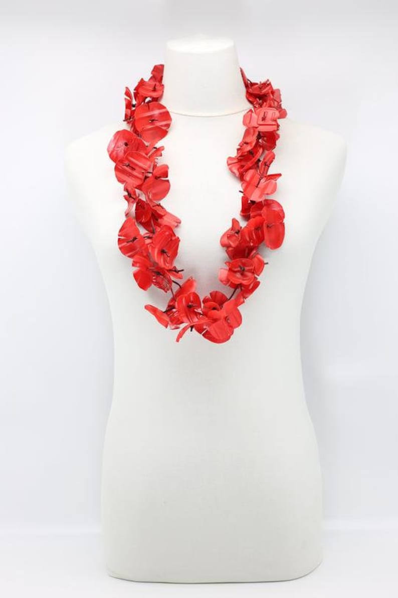 May include: A vibrant red floral necklace displayed on a white mannequin. The necklace is composed of numerous red flower-shaped elements, creating a layered and eye-catching design. The bright red color contrasts with the white background.