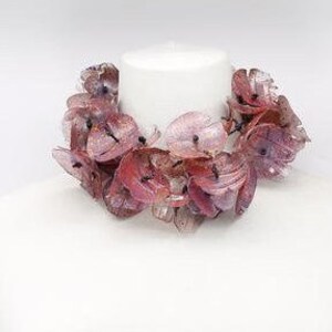 May include: A floral necklace with translucent, petal-shaped beads in shades of pink and red. The necklace is displayed on a white mannequin bust, showcasing its design and color variations. The beads appear to be made of a lightweight material.