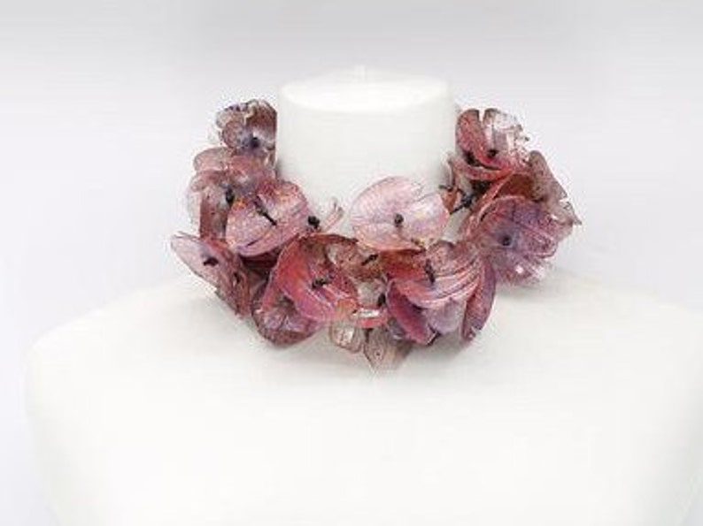 May include: A floral necklace with translucent, petal-shaped beads in shades of pink and red. The necklace is displayed on a white mannequin bust, showcasing its design and color variations. The beads appear to be made of a lightweight material.