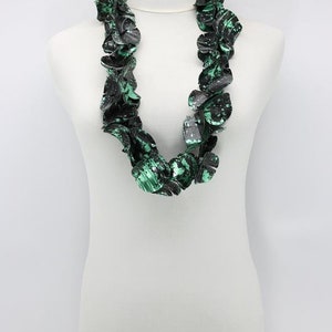 May include: A statement necklace displayed on a white mannequin. The necklace features large, irregular, petal-like shapes in black and green, creating a bold, textured look. The color palette is primarily black with splashes of vibrant green.