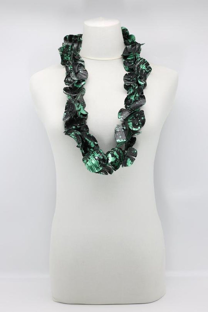 May include: A statement necklace displayed on a white mannequin. The necklace features large, irregular, petal-like shapes in black and green, creating a bold, textured look. The color palette is primarily black with splashes of vibrant green.