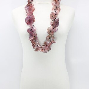 May include: A necklace made of translucent, petal-shaped beads in shades of pink and purple. The beads are strung together to form a statement piece, displayed on a white mannequin. The necklace has a delicate, floral appearance.