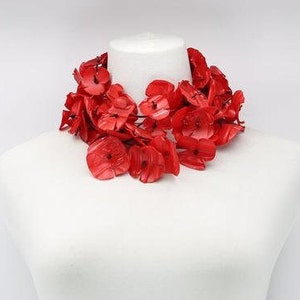 May include: A vibrant red floral necklace displayed on a white mannequin. The necklace features numerous flower-shaped beads, creating a bold and eye-catching accessory. The bright red color contrasts with the white background, highlighting the necklace's design.
