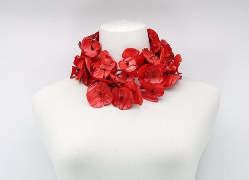May include: A vibrant red floral necklace displayed on a white mannequin. The necklace features numerous flower-shaped beads, creating a bold and eye-catching accessory. The bright red color contrasts with the white background, highlighting the necklace's design.