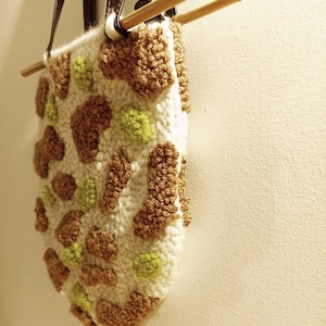 May include: A white crocheted bag with brown and green yarn pom-poms. The bag has a brown leather strap and is hanging from a wooden dowel.