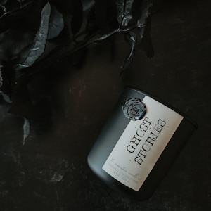 Puede incluir: Un tarro de vela negro con una etiqueta blanca que dice "Ghost Stories". El tarro tiene un sello de cera negro con un diseño de luna y estrellas. La vela es de 8 oz (226 g) y está hecha con cera de coco natural.