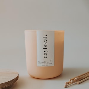 May include: A peach-colored candle in a glass jar with a white label that reads "daybreak." The label also states "the moonstone candle co." and "NATURAL COCONUT WAX 12 OZ / 340 G." A wooden lid and matches are visible.