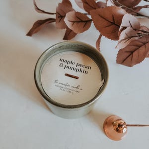 Könnte beinhalten: Eine Keramikkerze mit Holzdeckel, beschriftet mit "maple pecan & pumpkin". Die Kerze hat 283 g und besteht aus natürlichem Kokoswachs. Ein kupferner Kerzenlöscher befindet sich im Vordergrund, und Herbstlaub ist im Hintergrund.