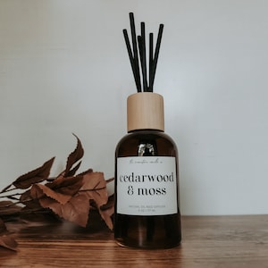 Puede incluir: Una botella de vidrio marrón con una tapa de madera y palitos difusores negros. La botella tiene una etiqueta blanca que dice "the mountain candle co. cedarwood & moss natural oil reed diffuser 6 oz / 177 ml".