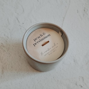 May include: A gray ceramic candle jar with a white lid. The lid has a label that reads "peach & persimmon the moonstone candle co NATURAL COCONUT WAX 10 OZ / 283 G".