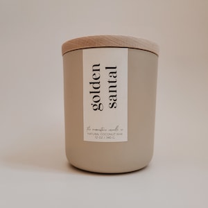 Puede incluir: Una vela beige con una tapa de madera y una etiqueta que dice "golden santal". La etiqueta también dice "the moonshine candle co. natural coconut wax 12 oz / 340 g".