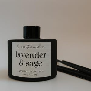 May include: A black, square-shaped oil diffuser bottle with a black cap. The label reads "lavender & sage" and "natural oil diffuser, 6 oz / 177 ml". Black diffuser sticks are visible to the right. The product is from "the moonstone candle co."