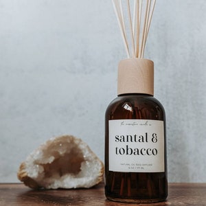 Puede incluir: Una botella de vidrio marrón con tapa de madera y palitos difusores. La etiqueta dice "santal & tobacco". Una geoda está a la izquierda de la botella. El difusor es de 177 ml.