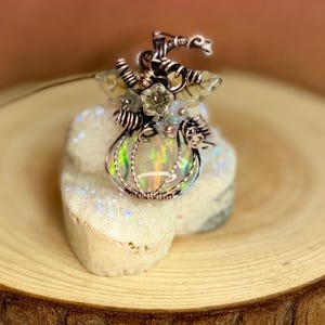 May include: A wire-wrapped pendant shaped like a pumpkin, featuring a central iridescent opal stone encased in copper wire. The design includes small metal flower accents and coiled wire tendrils at the top, creating a detailed botanical-inspired jewelry piece.