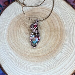 May include: A copper wire wrapped pendant featuring a colorful stone with a red gemstone accent. The pendant is hanging from a brown leather cord.