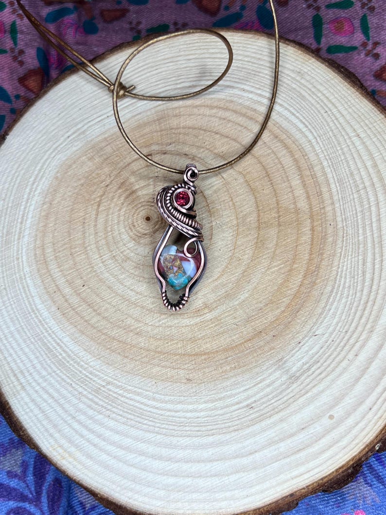 May include: A copper wire wrapped pendant featuring a colorful stone with a red gemstone accent. The pendant is hanging from a brown leather cord.