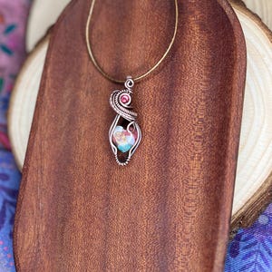May include: A delicate wire-wrapped pendant necklace featuring a pink and blue gemstone with a smaller pink gemstone accent. The necklace is displayed on a wooden surface.