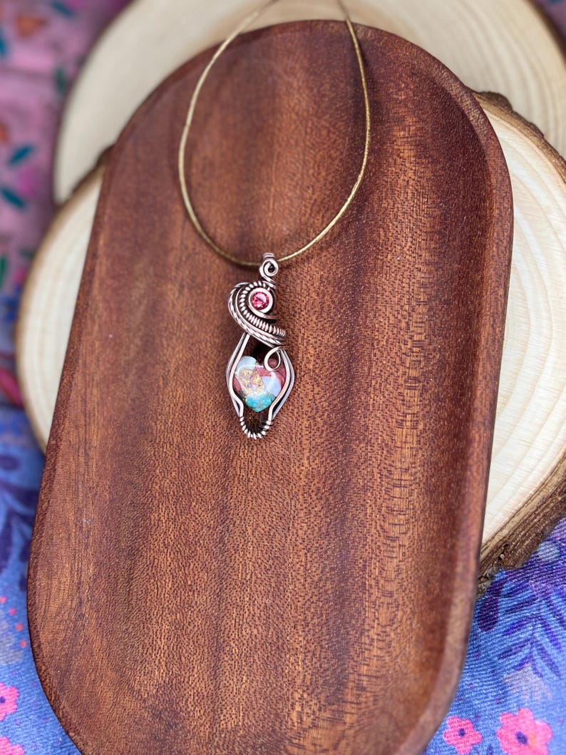 May include: A delicate wire-wrapped pendant necklace featuring a pink and blue gemstone with a smaller pink gemstone accent. The necklace is displayed on a wooden surface.