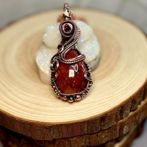 May include: A handcrafted copper pendant featuring a large, reddish-orange gemstone. The stone is intricately wrapped with copper wire, accented with small, dark red beads and a single, round, dark red stone at the top. The pendant is displayed on a wooden surface.