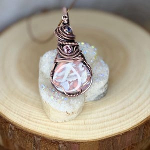 May include: A handcrafted pendant featuring a pink and white stone wrapped in copper wire. The wirework includes decorative coils and small, colorful gemstones. The pendant hangs from a thin copper chain, showcasing a unique, artistic design.