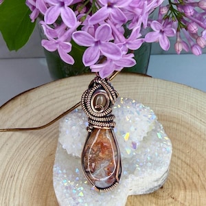 May include: A handcrafted copper wire-wrapped pendant with a teardrop-shaped agate stone. The stone features a mix of brown, cream, and orange hues. The pendant hangs on a thin copper chain, displayed on a white crystal cluster, with purple flowers in the background.