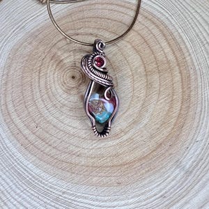 May include: A copper wire wrapped pendant featuring a multi-colored stone with a red gemstone accent. The pendant is hanging from a brown leather cord.
