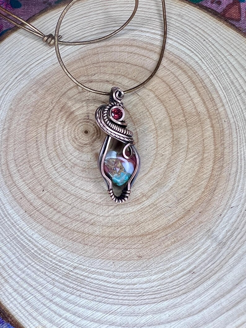 May include: A copper wire wrapped pendant featuring a multi-colored stone with a red gemstone accent. The pendant is hanging from a brown leather cord.