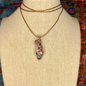 May include: A copper wire wrapped pendant necklace with a red gemstone and a multi-colored stone inside. The necklace is on a brown cord.