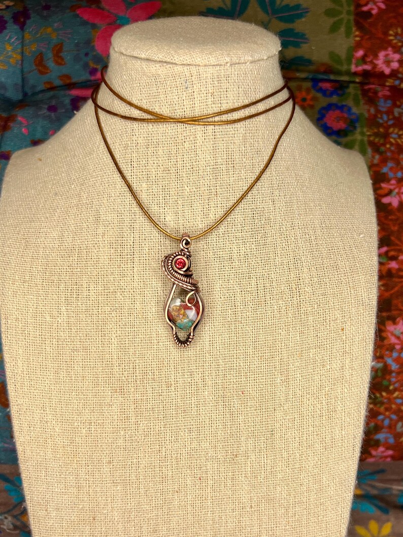 May include: A copper wire wrapped pendant necklace with a red gemstone and a multi-colored stone inside. The necklace is on a brown cord.