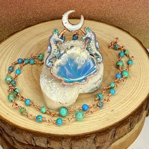 May include: A necklace featuring a large iridescent shell-shaped pendant topped with a crescent moon and two seahorses, all framed in copper wire. The chain consists of copper wire links interspersed with round turquoise and blue marbled beads.