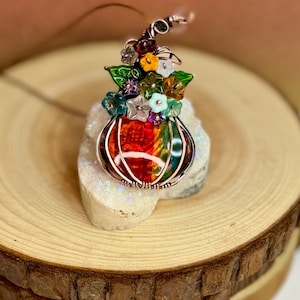 May include: A wire-wrapped pendant featuring a central iridescent pumpkin-shaped bead with rainbow colors. The top is adorned with a cluster of small, multi-colored glass flowers and green leaves in shades of yellow, white, blue, and purple.
