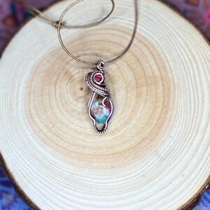 May include: A copper wire wrapped pendant necklace with a colorful stone and a small red gemstone. The necklace is on a wooden surface.