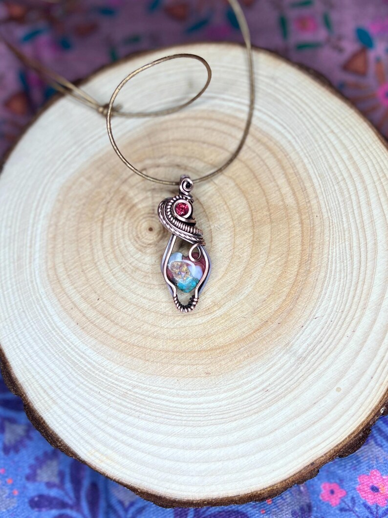 May include: A copper wire wrapped pendant necklace with a colorful stone and a small red gemstone. The necklace is on a wooden surface.