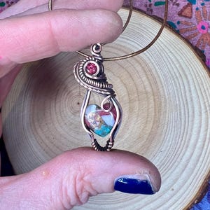 May include: A copper wire wrapped pendant featuring a heart-shaped stone with pink, white, and turquoise colors. A small red gemstone is incorporated into the wire wrapping.