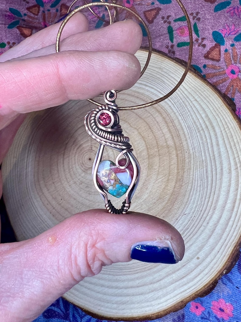 May include: A copper wire wrapped pendant featuring a heart-shaped stone with pink, white, and turquoise colors. A small red gemstone is incorporated into the wire wrapping.
