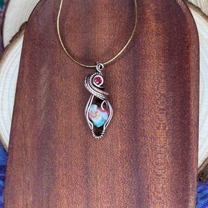 May include: A silver wire wrapped pendant necklace with a heart-shaped glass stone featuring a mix of blue, green, and pink colors. A small red gemstone is incorporated into the wire design.