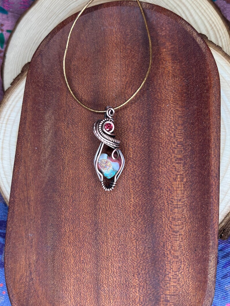 May include: A silver wire wrapped pendant necklace with a heart-shaped glass stone featuring a mix of blue, green, and pink colors. A small red gemstone is incorporated into the wire design.