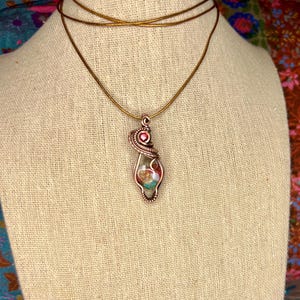 May include: A copper wire wrapped pendant necklace with a pink and green stone. The necklace is on a brown fabric mannequin.