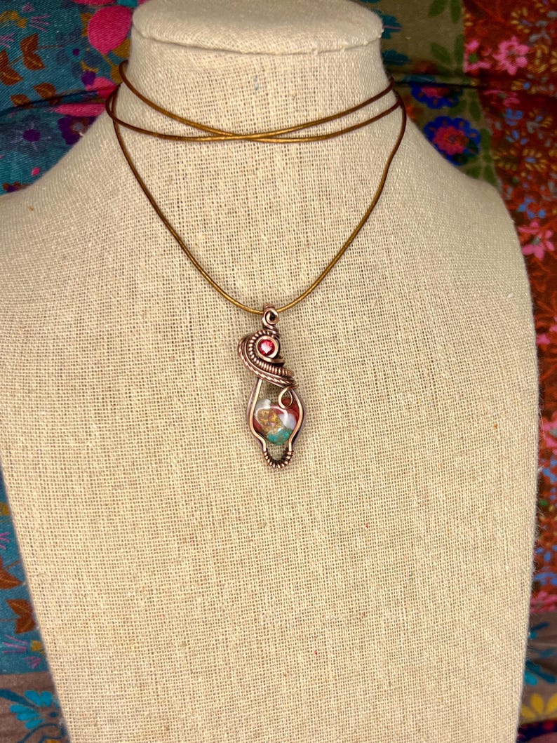 May include: A copper wire wrapped pendant necklace with a pink and green stone. The necklace is on a brown fabric mannequin.