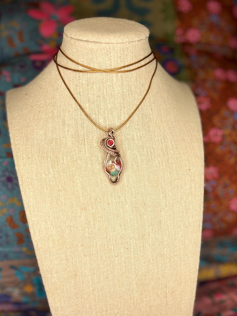 May include: A gold chain necklace with a pendant featuring a multi-colored stone wrapped in wire and a red gemstone accent.
