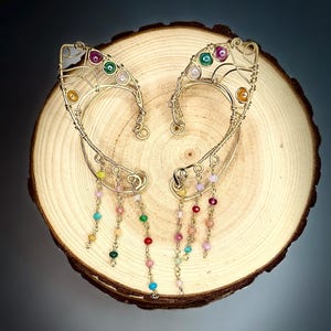 May include: A pair of gold-toned wire elf ear cuffs. Each cuff features colorful beads and dangling chains. The cuffs are adorned with various colored beads, including pink, green, yellow, and blue. They are displayed on a wooden surface.
