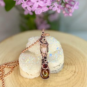 May include: A copper wire-wrapped pendant necklace featuring a teardrop-shaped, multi-colored stone. The pendant has a pink, oval-shaped stone and a red, round stone. The necklace is displayed on a white, crystal-like surface with purple flowers in the background.