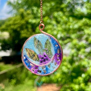 May include: A round, copper-colored pendant with a blue and purple design. The pendant features a central arrangement of green and purple elements, encased in a clear resin. It hangs from a delicate copper chain, with a blurred green background.