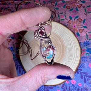 May include: A copper wire wrapped pendant featuring a heart-shaped stone with pink, white, and brown swirls. A small, dark red gemstone is incorporated into the wire wrapping.