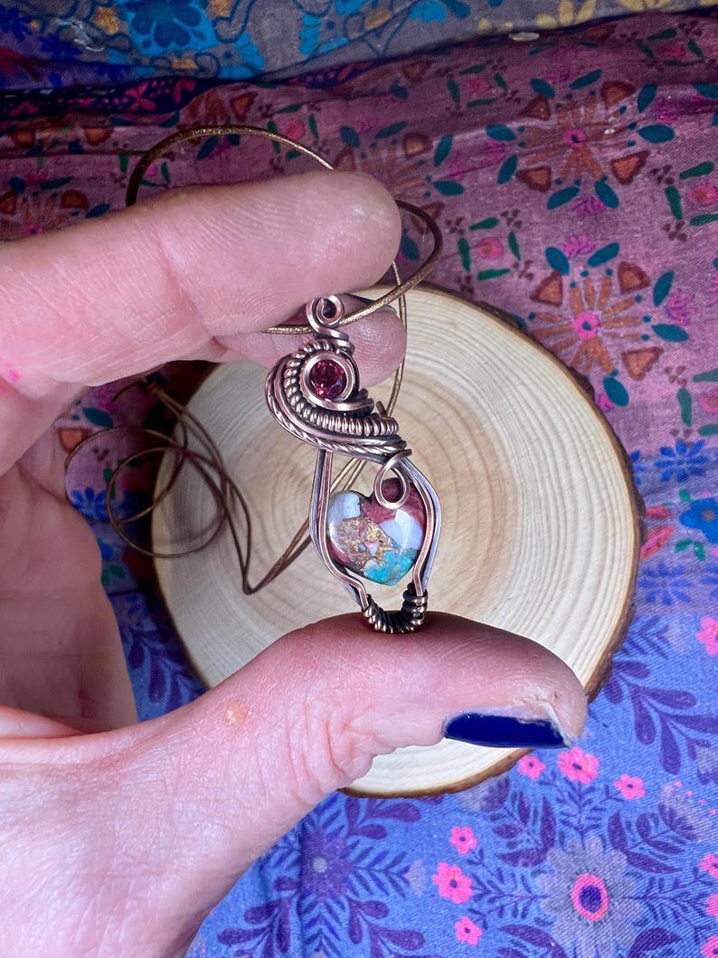 May include: A copper wire wrapped pendant featuring a heart-shaped stone with pink, white, and brown swirls. A small, dark red gemstone is incorporated into the wire wrapping.