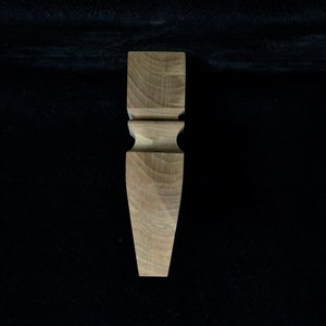 Könnte beinhalten: Ein Möbelbein aus Holz mit gedrehtem Design. Das Bein ist hellbraun und hat eine quadratische Oberseite und eine spitze Unterseite.