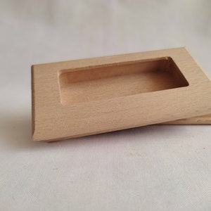 Op de afbeelding: Een kleine, rechthoekige houten doos met een verzonken rechthoekige opening. De doos is gemaakt van lichtgekleurd hout en heeft een licht schuine bovenkant. De doos wordt waarschijnlijk gebruikt voor opslag of weergave.