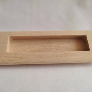 May include: A rectangular wooden drawer pull with a recessed handle. The pull is made of light-colored wood and has a smooth finish.