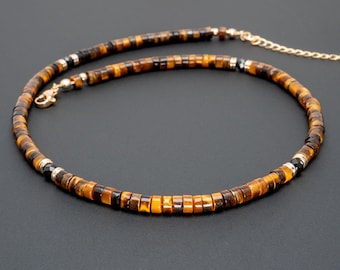May include: A beaded necklace featuring tiger's eye stones in shades of brown and gold. The necklace has a gold-colored chain and clasp. The beads are small and square-shaped, creating a textured look. The necklace is displayed on a dark background.